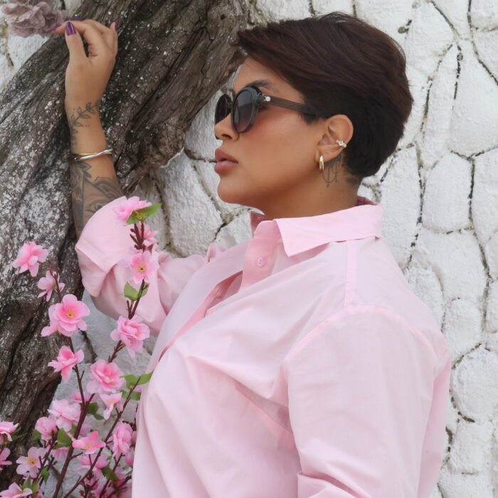 Mulher de perfil usando óculos de sol Blue Puya cinza com armação arredondada, lentes degradê e camisa rosa claro, encostada em uma árvore com flores cor-de-rosa.