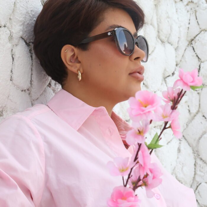 Mulher usando óculos de sol Blue Puya cinza com lentes degradê, camisa rosa e segurando um galho de flores cor-de-rosa, em frente a uma parede de pedras brancas.