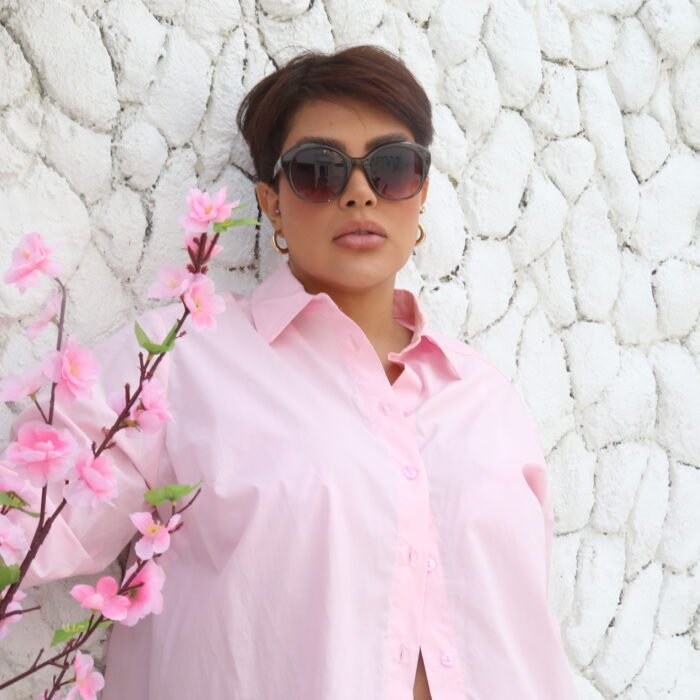 Mulher usando óculos de sol Blue Puya cinza com lentes degradê e camisa rosa claro, posando em frente a uma parede de pedras brancas com galhos de flores cor-de-rosa.