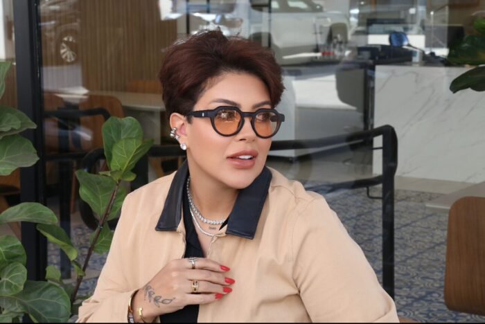 Mulher elegante usando óculos de sol Titan Arum Preto da Use Let’s Brasil com lentes alaranjadas, blazer bege com detalhes pretos, sentada em uma cafeteria moderna com plantas ao fundo
