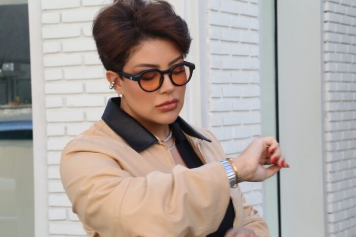 Mulher estilosa usando óculos de sol Titan Arum Preto da Use Let’s Brasil, com lentes alaranjadas, blazer bege e camisa preta, olhando para o relógio em ambiente urbano