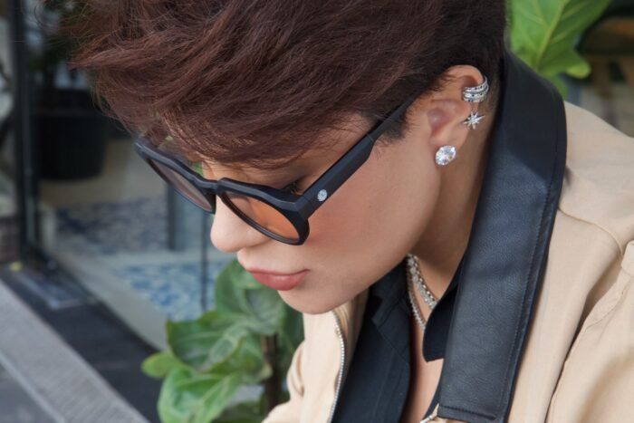 Mulher estilosa usando óculos de sol Titan Arum Preto da Use Let’s Brasil com lentes alaranjadas, blazer bege com gola preta, olhando para baixo em um ambiente externo com plantas