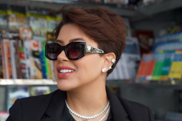 Mulher sorrindo, usando óculos de sol Black Bat Flower da Use Let’s, com armação preta robusta e lentes escuras, em frente a uma banca de revistas