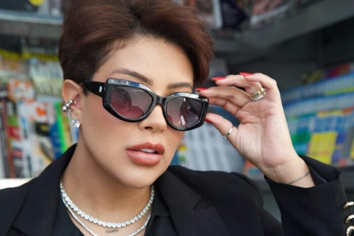Mulher estilosa usando óculos de sol Black Bat Flower da Use Let’s, com armação preta e lentes escuras, em cenário urbano com banca de revistas ao fundo