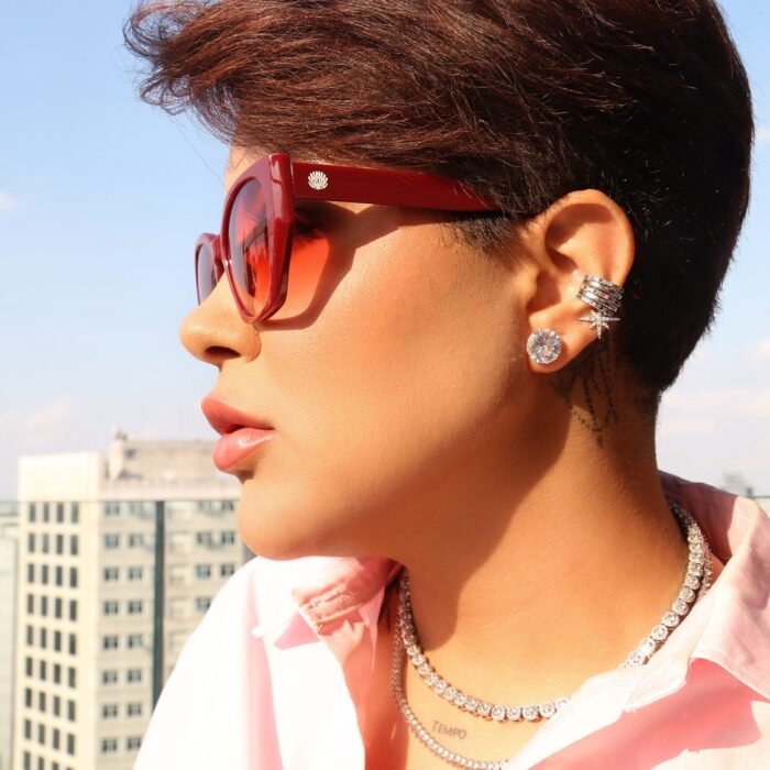 Mulher estilosa usando óculos de sol vermelho modelo Hooker’s Lips da Use Let’s Brasil, de perfil, com cabelo curto e camisa rosa, posando em um rooftop urbano com prédios ao fundo