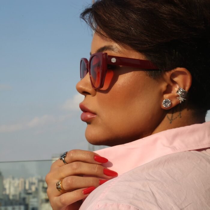 Mulher estilosa usando óculos de sol vermelho modelo Hooker’s Lips da Use Let’s Brasil, de perfil, com camisa rosa e unhas vermelhas, segurando a gola da camisa em um rooftop com prédios ao fundo