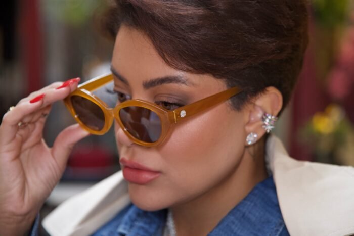 Mulher elegante ajustando óculos de sol Fire Lily da Use Let’s Brasil, modelo em acetato caramelo moderno com lentes marrons, look casual com camisa jeans e blazer claro