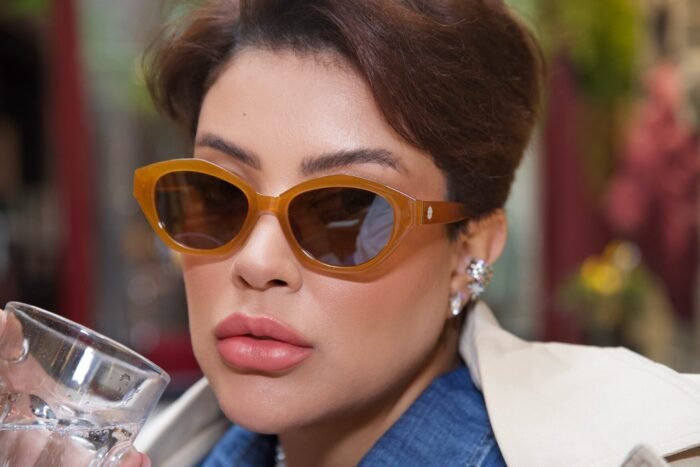 Mulher estilosa usando óculos de sol Fire Lily da Use Let’s Brasil na cor caramelo moderno, segurando uma taça, com look casual elegante e maquiagem impecável