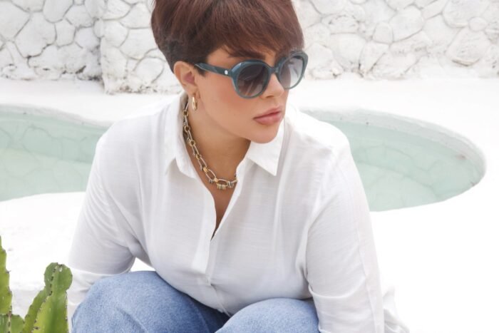 Mulher usando óculos de sol Blue Puya azul, com armação arredondada e lentes degradê, sentada em ambiente externo de tom branco, com parede de pedras e vegetação ao fundo.
