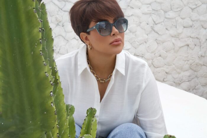 Mulher estilosa usando óculos de sol Blue Puya da Use Let’s Brasil, cor azul petróleo, camisa branca e jeans, sentada em ambiente externo com parede branca e planta ao fundo