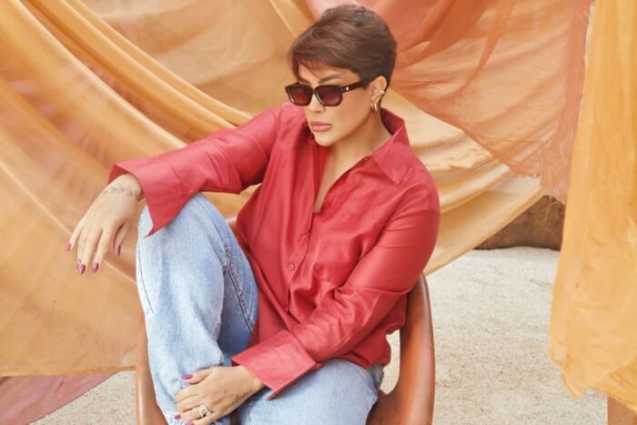 Mulher estilosa usando óculos de sol Desert Rose Marrom da Use Let’s Brasil, com armação marrom e lentes marrom degradê, sentada em cadeira, vestindo camisa vermelha e jeans, em ambiente com tecidos em tons terrosos.