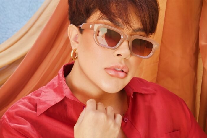 Close de mulher usando óculos de sol Desert Rose Nude da Use Let’s Brasil, com camisa vermelha, fundo com tecidos em tons terrosos e azul