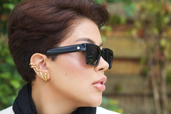 Perfil de uma mulher estilosa usando óculos de sol preto modelo Martagon da Use Let’s Brasil, com lentes escuras e detalhe metálico na haste, combinando com brincos dourados, em ambiente externo com vegetação desfocada ao fundo