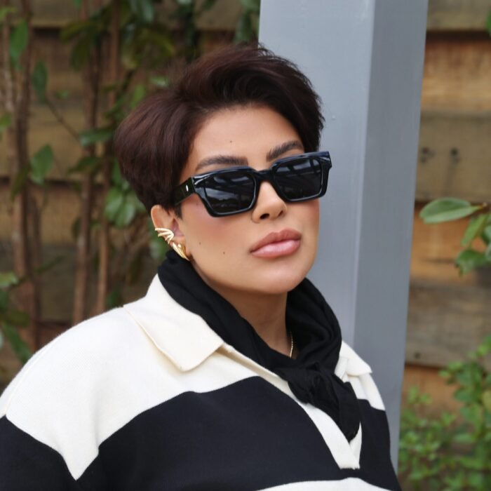 Mulher estilosa usando óculos de sol preto modelo Martagon da Use Let’s Brasil, com lentes escuras, brincos dourados e look moderno em preto e branco, posando em ambiente externo com plantas ao fundo