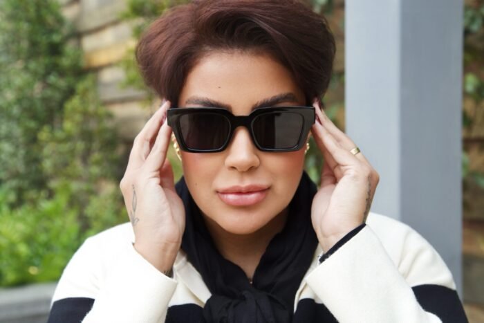 Mulher estilosa usando óculos de sol preto modelo Martagon da Use Let’s Brasil, segurando as hastes do óculos com as duas mãos, usando look preto e branco, em ambiente externo com vegetação desfocada ao fundo