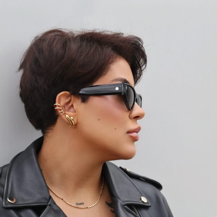 Vista lateral de uma mulher estilosa usando óculos de sol preto modelo Lilium da Use Let’s Brasil, com armação em acetato preto, lentes escuras e detalhe metálico na haste, usando jaqueta de couro preta e brincos dourados, posando em frente a uma parede cinza