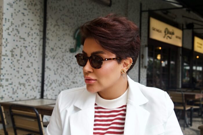 Mulher estilosa usando óculos de sol tartaruga modelo Calla da Use Let’s Brasil, com look elegante de blazer branco e blusa listrada em vermelho e branco, sentada em cafeteria de ambiente moderno