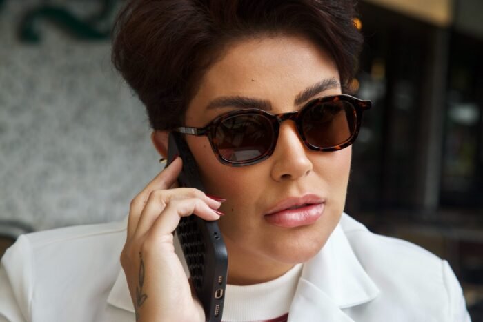Mulher estilosa usando óculos de sol tartaruga modelo Calla da Use Let’s Brasil, segurando um telefone, com look elegante composto por blazer branco e blusa listrada, em ambiente urbano de cafeteria